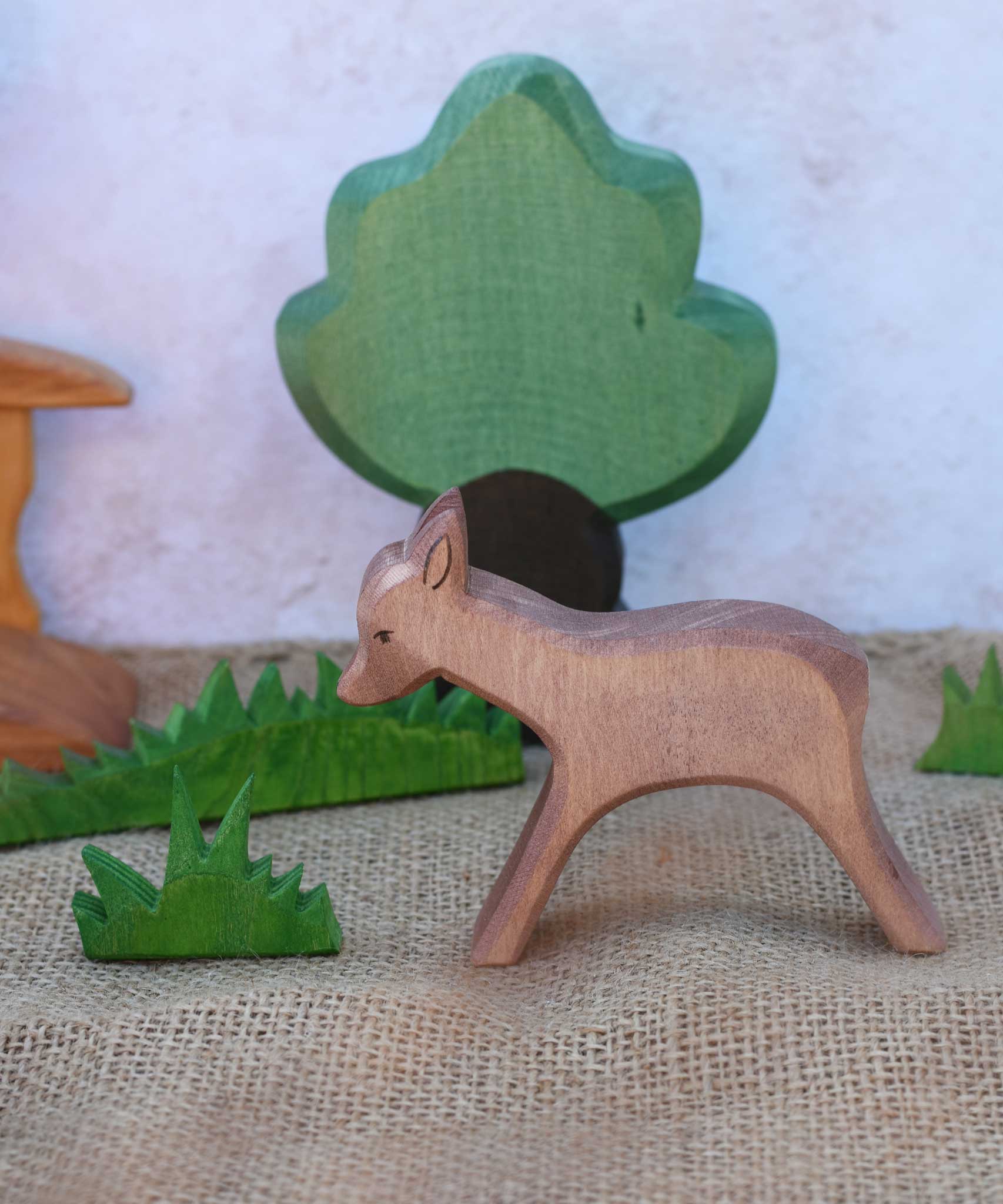 An Ostheimer wooden standing deer figure shown in a small world play scene set up with grass, oak tree and a bridge in the background.