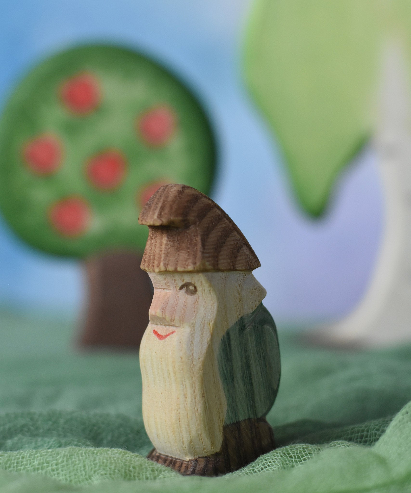 An Ostheimer standing fire tree dwarf wooden toy figure placed in a small world play scene. The dwarf figure has a green coloured outfit with brown hat and a long white beard. The figure has been placed on a green coloured Ostheimer play scarf. This wooden figure is part of a large range of expertly hand crafted, Waldorf inspired, wooden toys here at Babipur. 