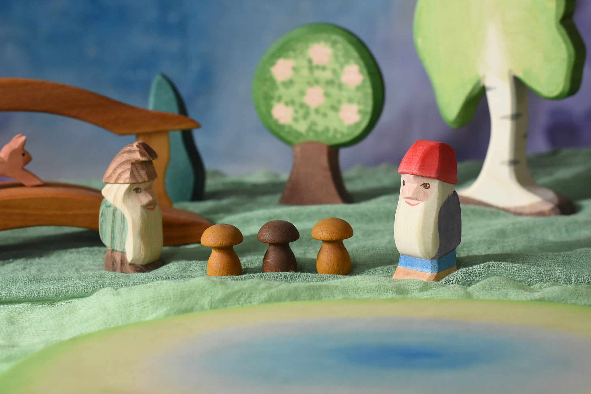 An Ostheimer standing fir tree and Bodo dwarf standing in front of an Ostheimer pond play piece in a small world play scene. There are wooden toy scenery pieces in the background including a apple tree and large birch tree. These wooden figures are part of a wide range of expertly hand crafted wooden toys here at Babipur. 
