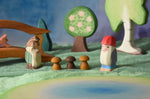 An Ostheimer standing fir tree and Bodo dwarf standing in front of an Ostheimer pond play piece in a small world play scene. There are wooden toy scenery pieces in the background including a apple tree and large birch tree. These wooden figures are part of a wide range of expertly hand crafted wooden toys here at Babipur. 
