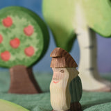 An Ostheimer standing fire tree dwarf wooden toy figure placed in a small world play scene. The dwarf figure has a green coloured outfit with brown hat and a long white beard. The figure has been placed on a green coloured Ostheimer play scarf. This wooden figure is part of a large range of expertly hand crafted, Waldorf inspired, wooden toys here at Babipur. 