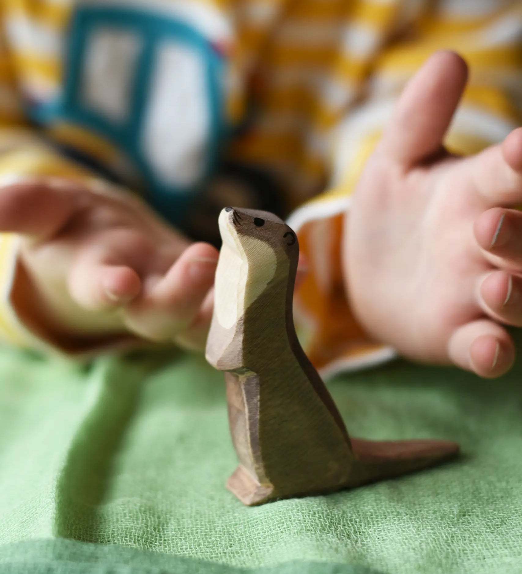 Ostheimer standing sea otter placed on a green cotton play cloth with a child's hand hovering each side of the figure.