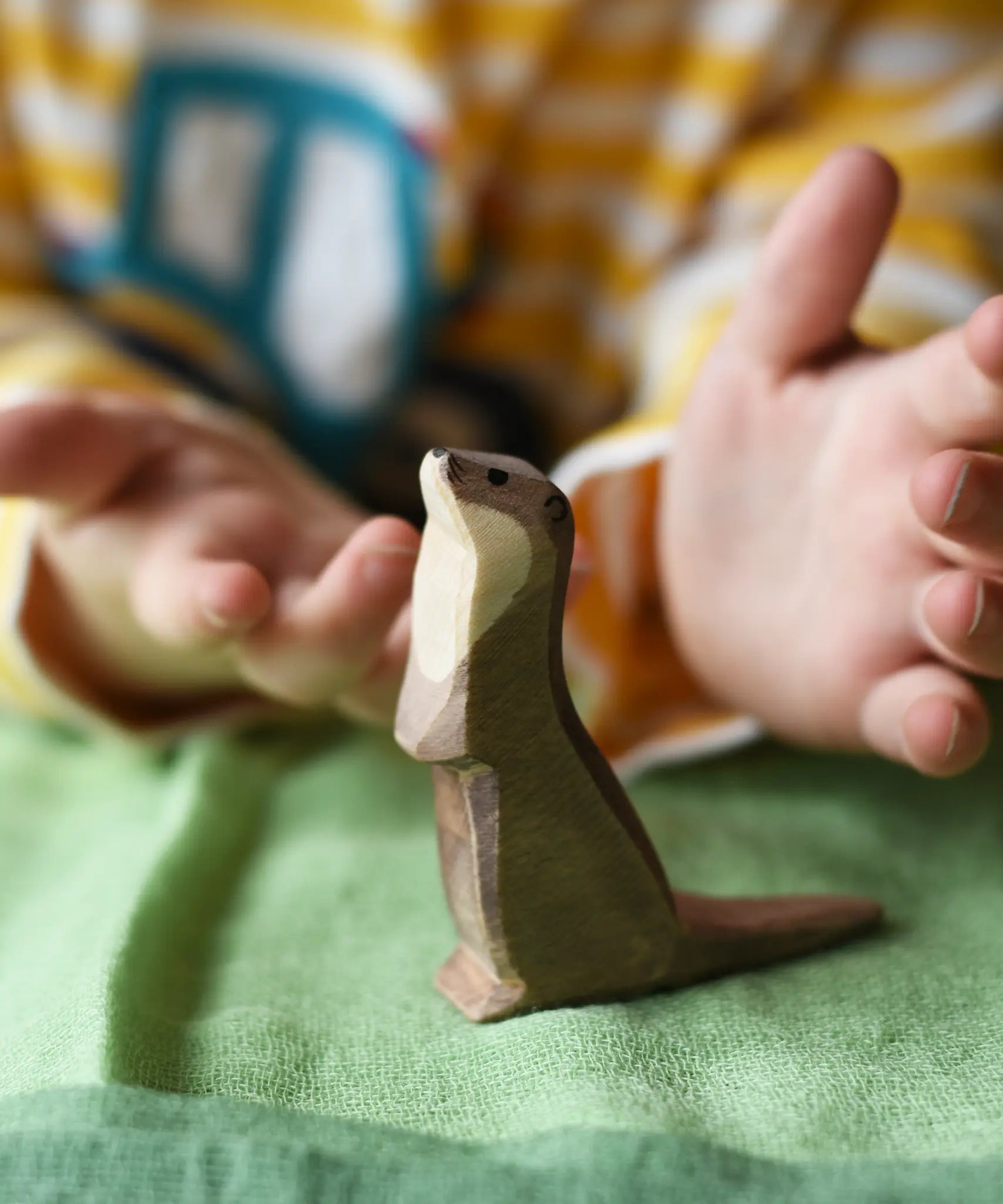 Ostheimer standing sea otter placed on a green cotton play cloth with a child's hand hovering each side of the figure.