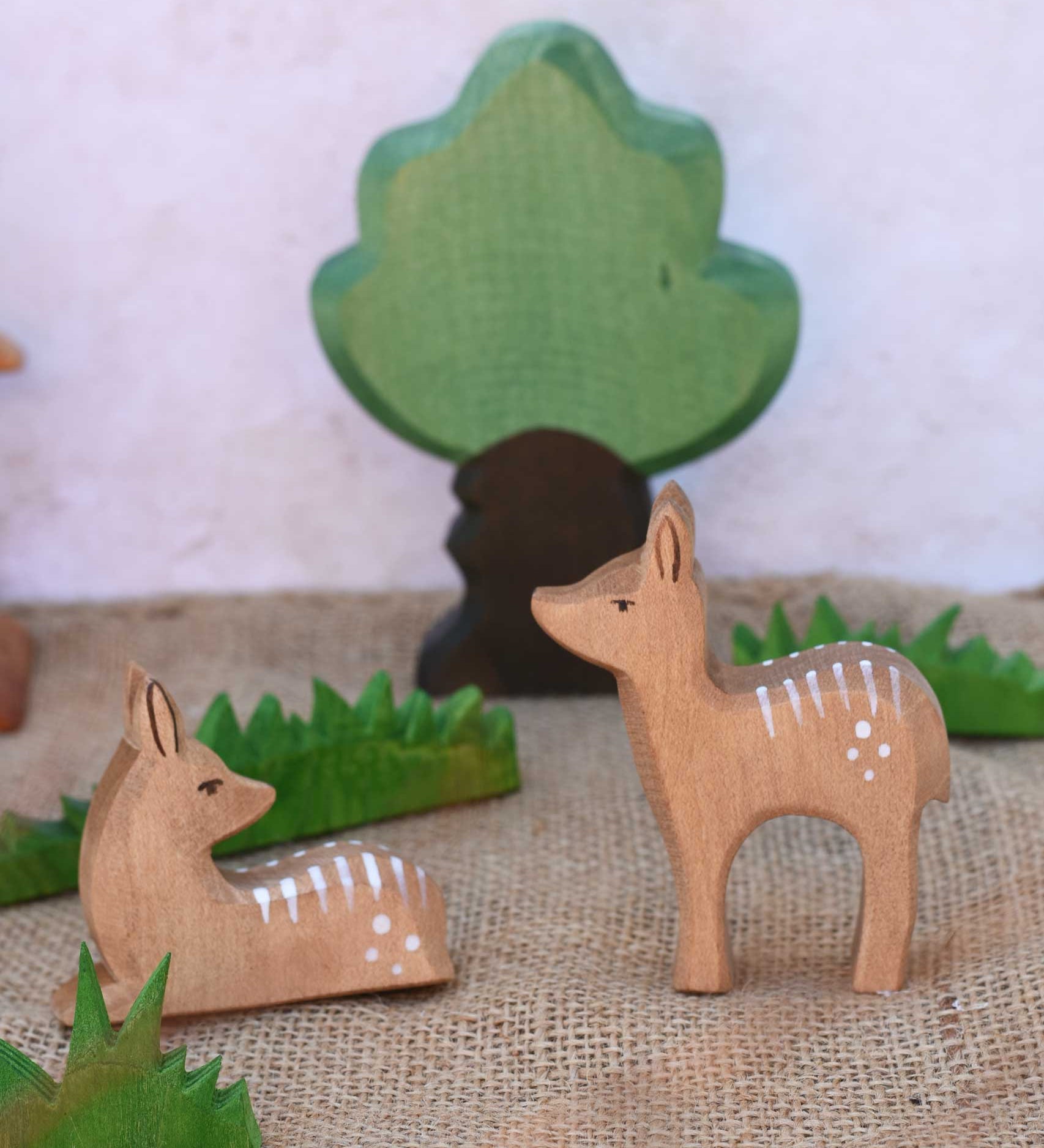 Ostheimer sitting and standing deer fawn wooden figures placed in a small world play scene with other wooden Ostheimer figure including wooden green grass and an oak tree.