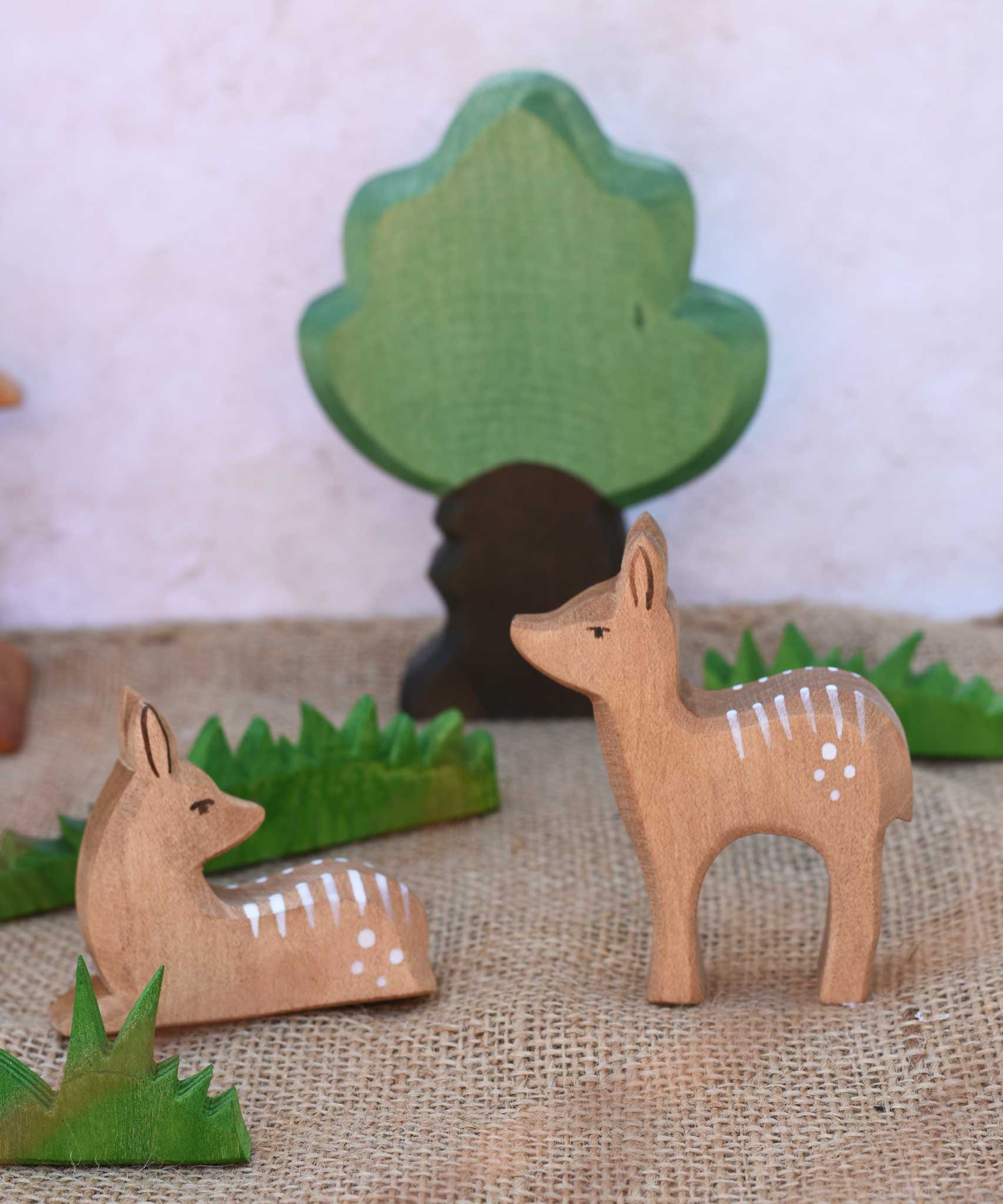 Ostheimer sitting and standing deer fawn wooden figures placed in a small world play scene with other wooden Ostheimer figure including wooden green grass and an oak tree.