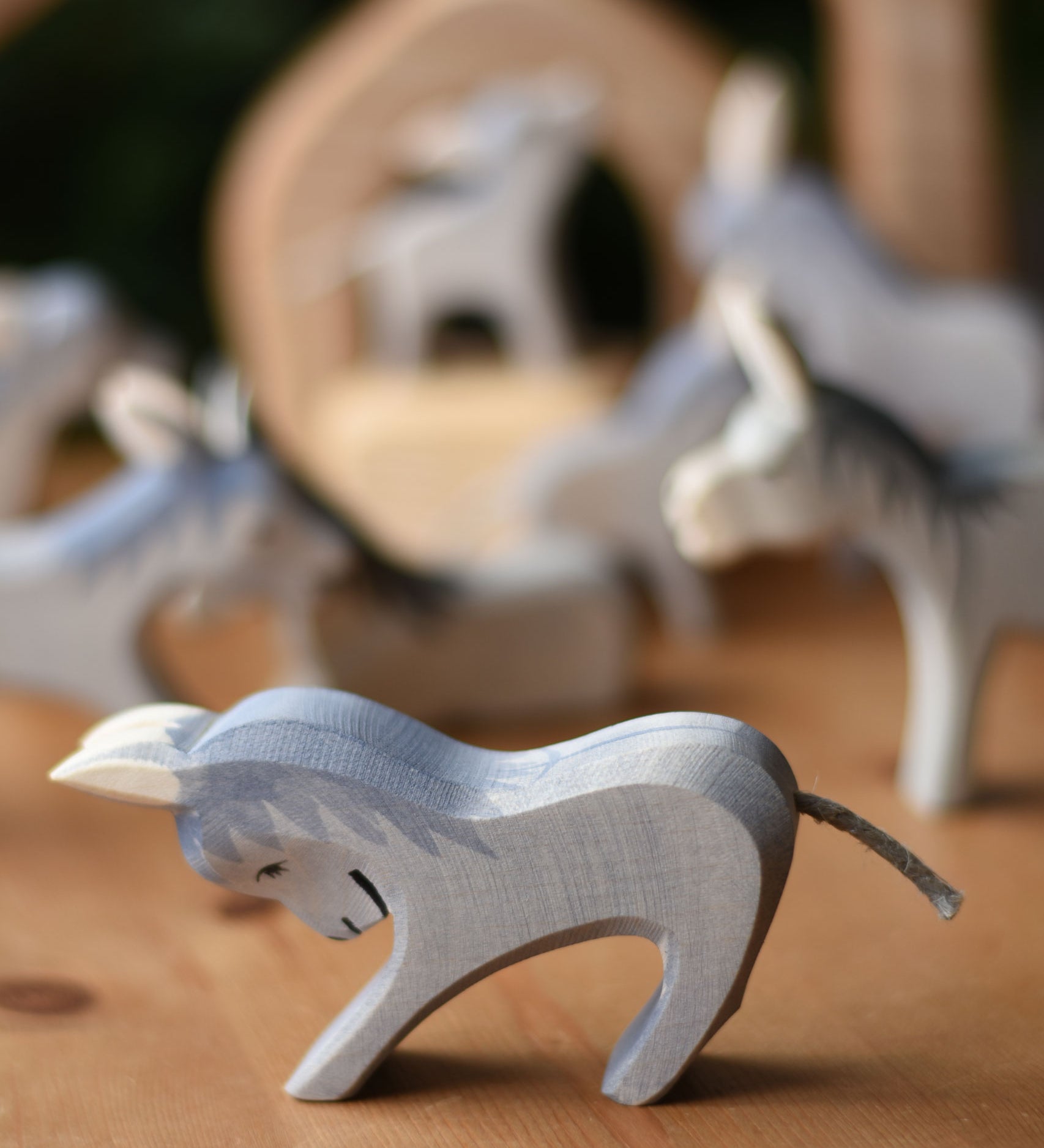 An Ostheimer stubborn donkey wooden toy figure placed on a wooden surface with other donkey's blurred in the background.  Ostheimer's range of hand crafted, heirloom quality wooden toys are available to buy at Babipur.