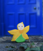 An Ostheimer Sunflower Flower Child placed on a stone wall outdoors in front of a blue door. This wooden figure is part of a large range of expertly hand crafted, Waldorf inspired, wooden toys here at Babipur. 