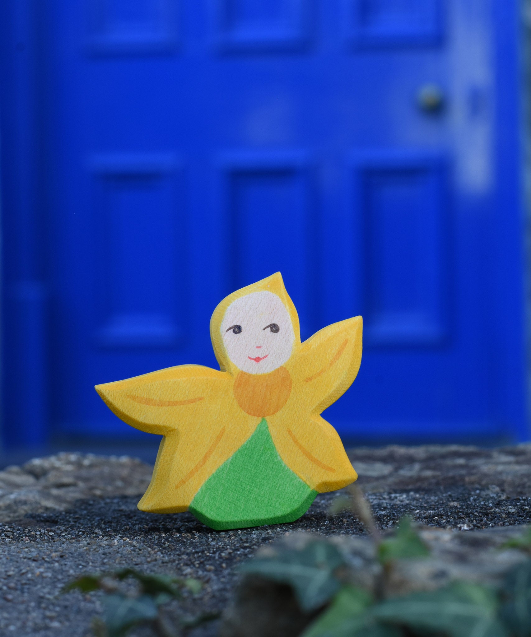 An Ostheimer Sunflower Flower Child placed on a stone wall outdoors in front of a blue door. This wooden figure is part of a large range of expertly hand crafted, Waldorf inspired, wooden toys here at Babipur. 