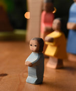 An Ostheimer toddler wooden toy figure with brown skin placed at the front of a small world play scene. These wooden figures are part of a wide range of expertly hand crafted wooden toys here at Babipur.