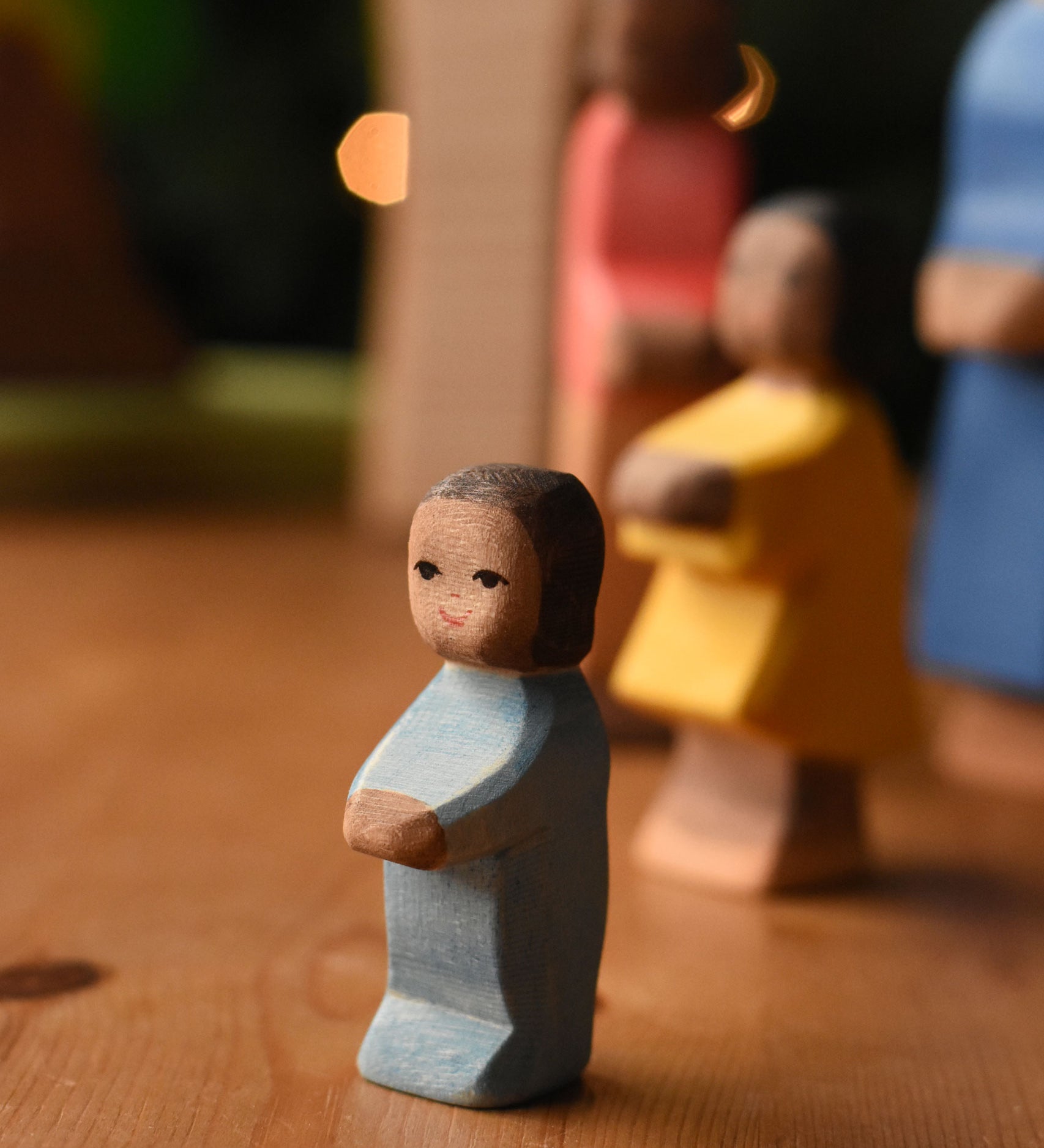 An Ostheimer toddler wooden toy figure with brown skin placed at the front of a small world play scene. These wooden figures are part of a wide range of expertly hand crafted wooden toys here at Babipur.