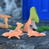 A family of four Ostheimer wooden Fox figures shown in a small world play scene set up on a wooden wall outdoors. The Ostheimer birch tree, grass and bridge with railing can be seen on the background. Check out our full range of expertly crafted wooden toy figures and scenery here at Babipur.