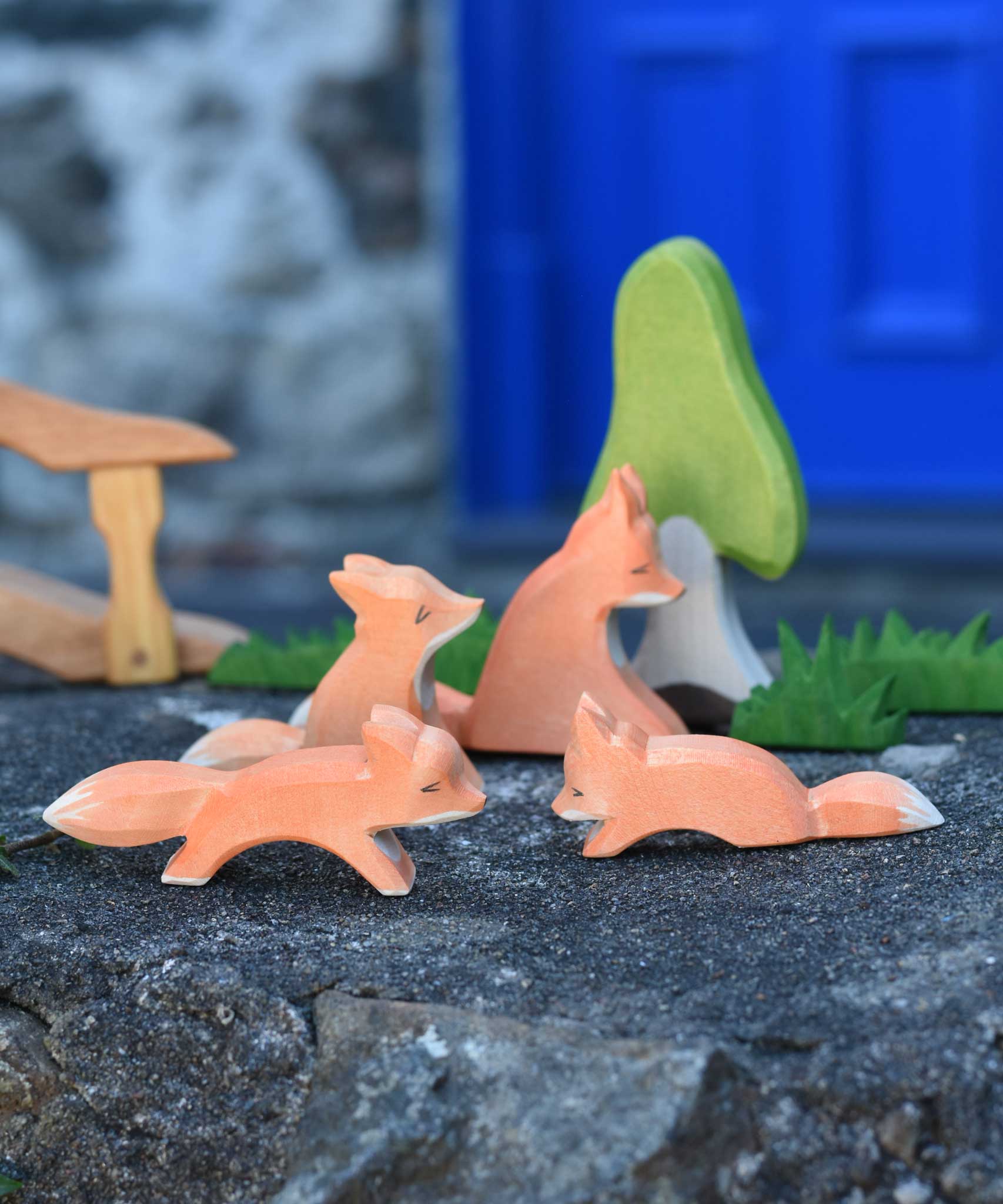 A family of four Ostheimer wooden Fox figures shown in a small world play scene set up on a wooden wall outdoors. The Ostheimer birch tree, grass and bridge with railing can be seen on the background. Check out our full range of expertly crafted wooden toy figures and scenery here at Babipur.