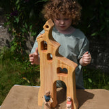 A child smiling standing behind an Ostheimer wooden bell tower. The child is reaching up for the bell. These wooden figures are part of a large range of expertly hand crafted, Waldorf inspired, wooden toys here at Babipur.