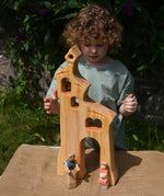 A child smiling standing behind an Ostheimer wooden bell tower. The child is reaching up for the bell. These wooden figures are part of a large range of expertly hand crafted, Waldorf inspired, wooden toys here at Babipur.