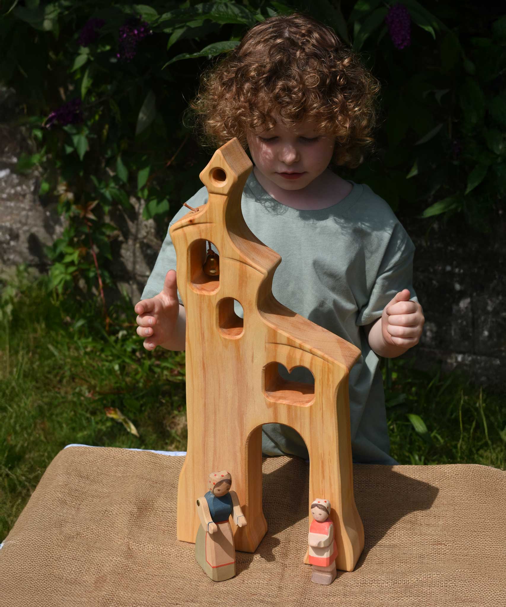 A child smiling standing behind an Ostheimer wooden bell tower. The child is reaching up for the bell. These wooden figures are part of a large range of expertly hand crafted, Waldorf inspired, wooden toys here at Babipur.