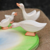 Ostheimer white goose with low neck wooden toy figure placed on an Ostheimer pond scene piece. Other wooden geese figures can be seen in the background. 