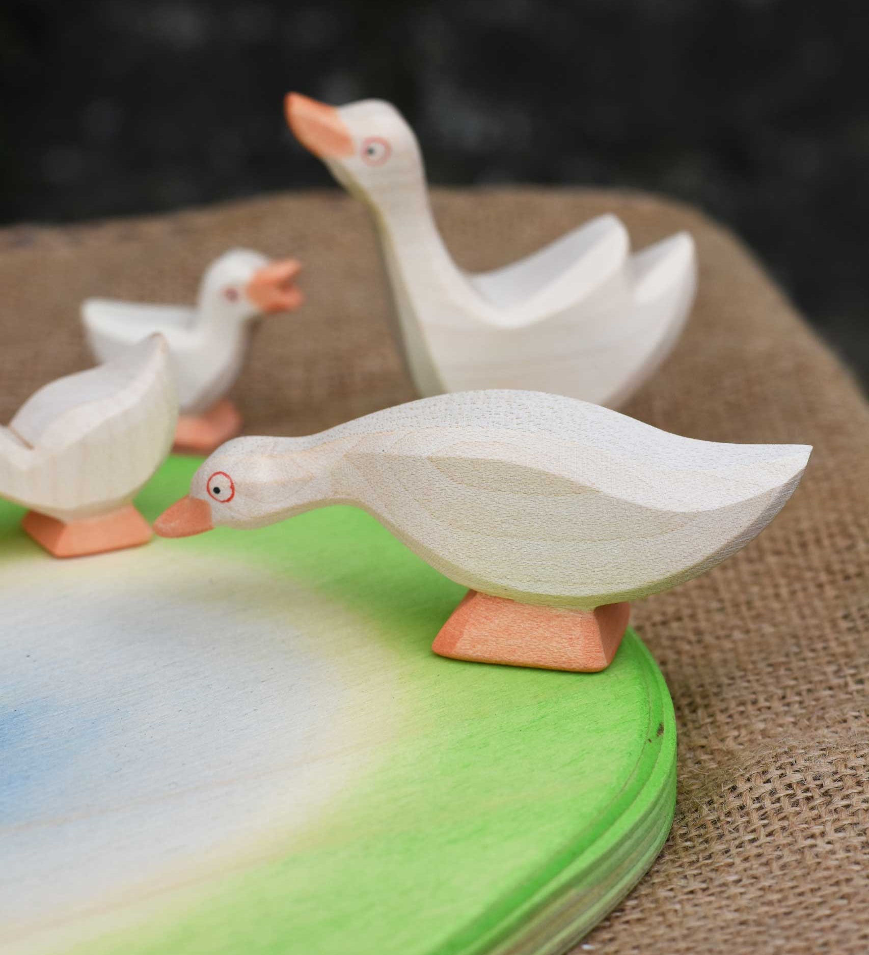 Ostheimer white goose with low neck wooden toy figure placed on an Ostheimer pond scene piece. Other wooden geese figures can be seen in the background. 