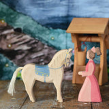 An Ostheimer hand made wooden Lady-In-Waiting toy figure and white horse with saddle placed on a wooden surface in a small world play scene. Other wooden toys including an Ostheimer water well can be seen in the background. These wooden figures are part of a wide range of expertly hand crafted wooden toys here at Babipur. 
