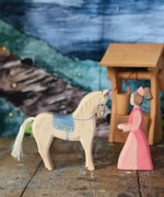 An Ostheimer hand made wooden Lady-In-Waiting toy figure and white horse with saddle placed on a wooden surface in a small world play scene. Other wooden toys including an Ostheimer water well can be seen in the background. These wooden figures are part of a wide range of expertly hand crafted wooden toys here at Babipur. 