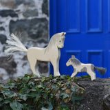 An Ostheimer white horse and pony figure placed on a stone wall outdoors. These wooden figures are part of a large range of expertly hand crafted, Waldorf inspired, wooden toys here at Babipur. 
