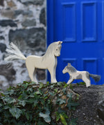 An Ostheimer white horse and pony figure placed on a stone wall outdoors. These wooden figures are part of a large range of expertly hand crafted, Waldorf inspired, wooden toys here at Babipur. 
