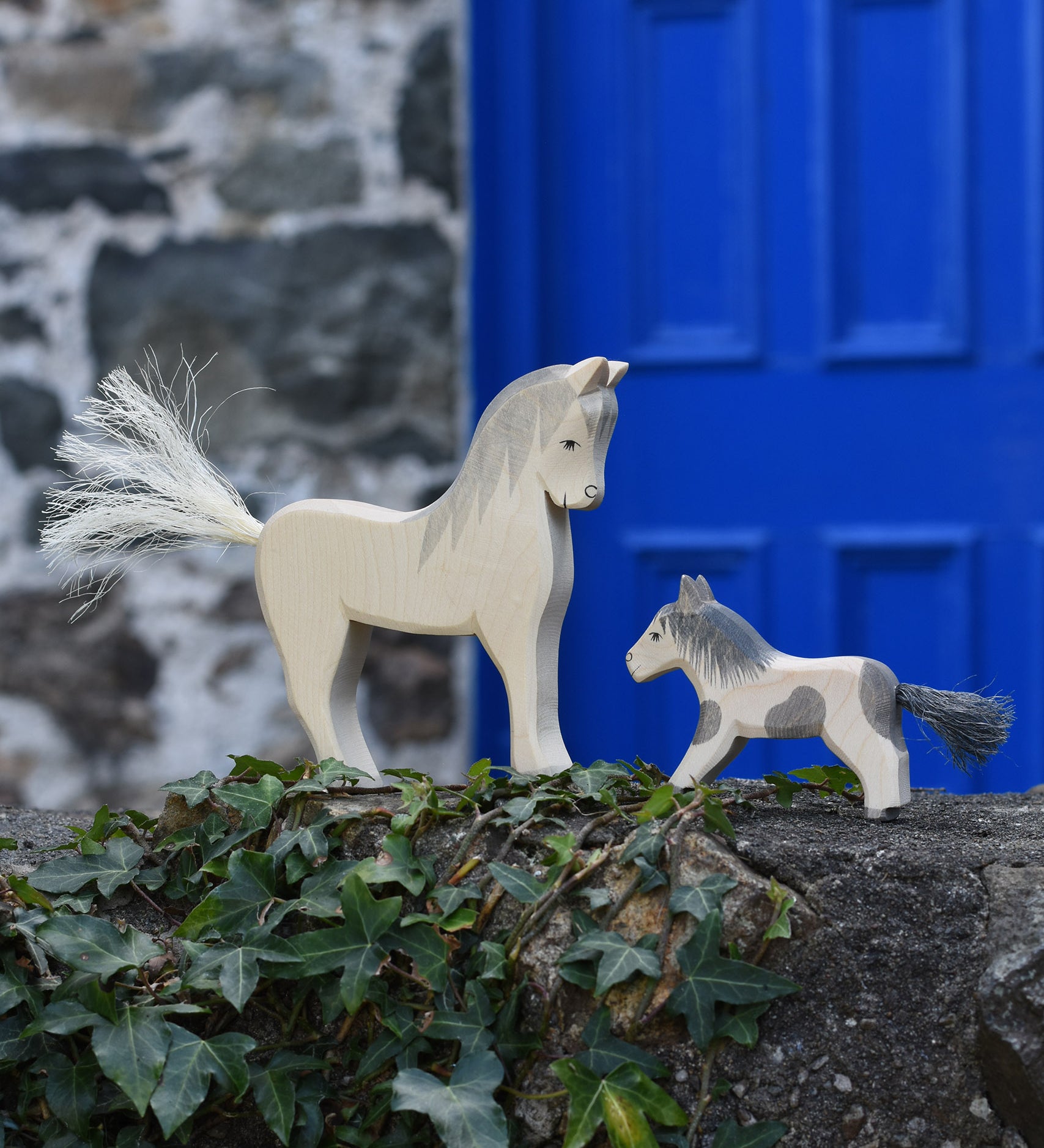 An Ostheimer white horse and pony figure placed on a stone wall outdoors. These wooden figures are part of a large range of expertly hand crafted, Waldorf inspired, wooden toys here at Babipur. 
