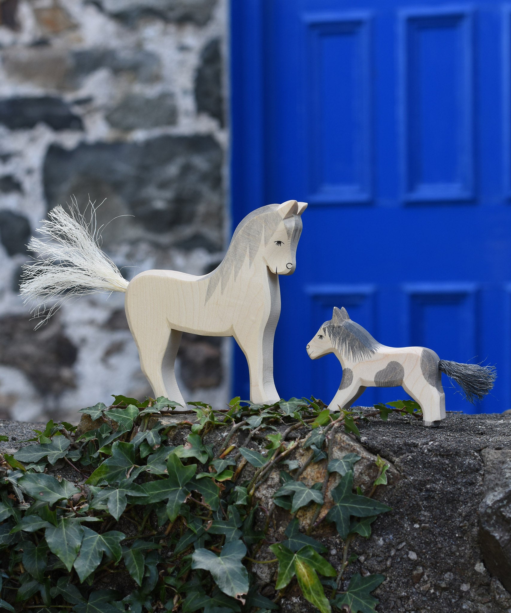 An Ostheimer white horse and pony figure placed on a stone wall outdoors. These wooden figures are part of a large range of expertly hand crafted, Waldorf inspired, wooden toys here at Babipur. 

