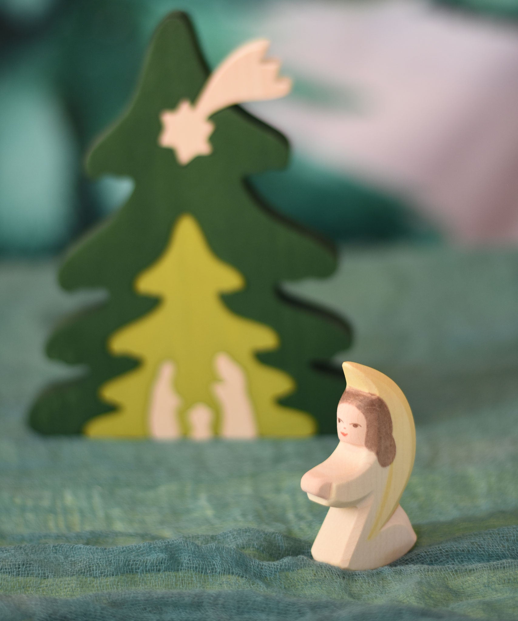 An Ostheimer little angel figure with white outfit placed at the front of small world play scene. There is a Bumbu nativity scene tree in the background. These wooden figures are part of a wide range of expertly hand crafted wooden toys here at Babipur.