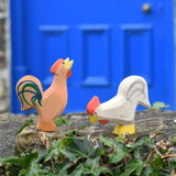 Ostheimer brown bremer rooster next to a white rooster wooden toy figure placed on a stone wall outdoors. 