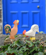Ostheimer brown bremer rooster next to a white rooster wooden toy figure placed on a stone wall outdoors. 