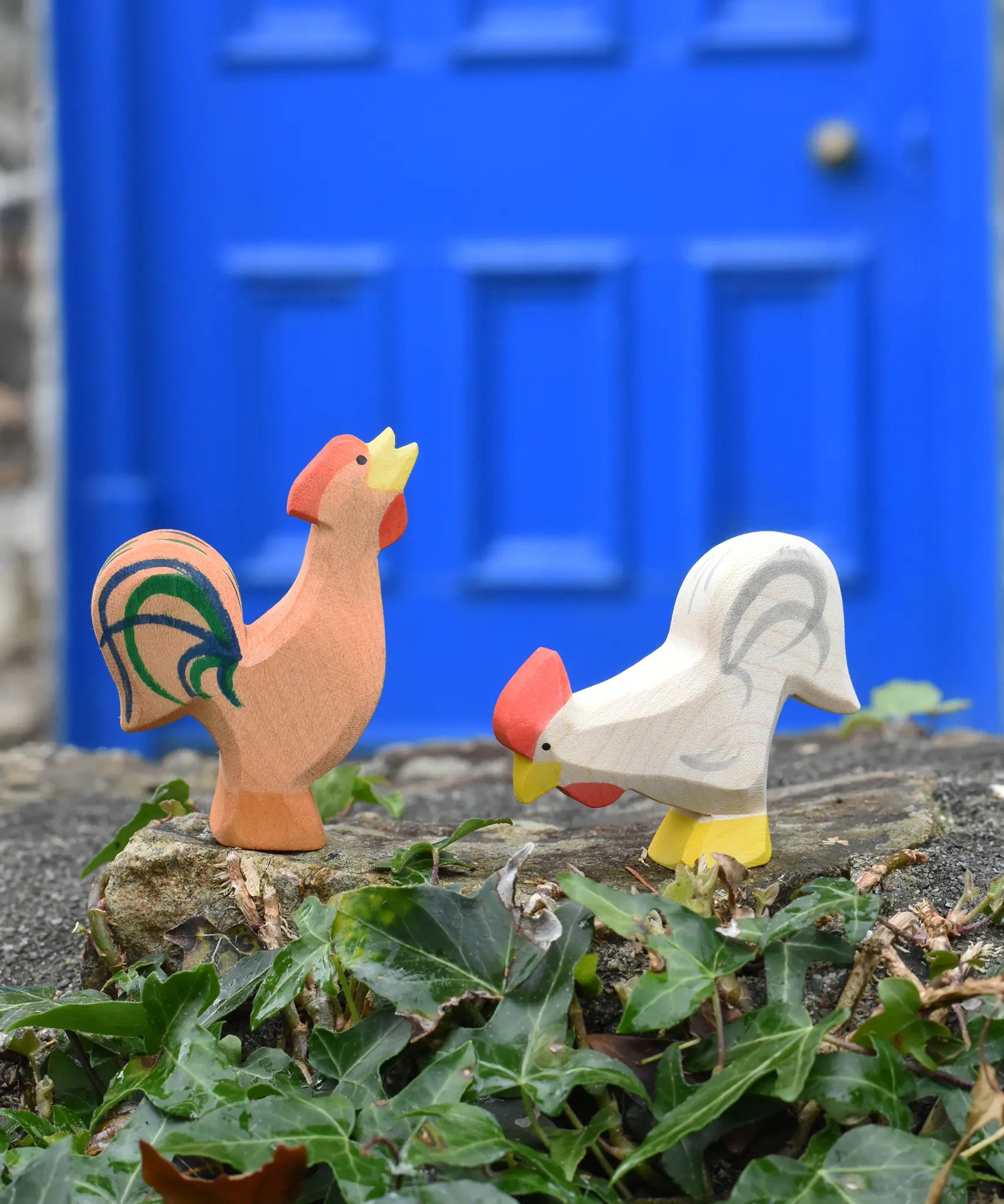 Ostheimer brown bremer rooster next to a white rooster wooden toy figure placed on a stone wall outdoors. 
