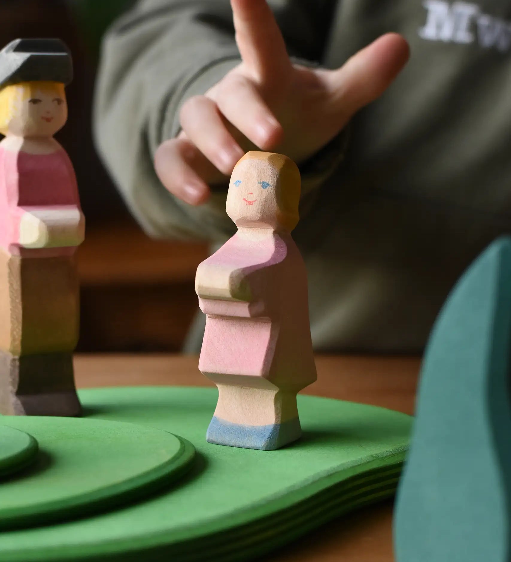 Ostheimer daughter wooden toy figure placed on green meadow grass play base. A child's hand an be seen reaching for teh girl figure in the backround.