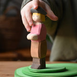 Child's hand reaching for the Ostheimer son wooden toy figure. The handmade boy figure is wearing a black hat, red top, brown trousers and brown boots.