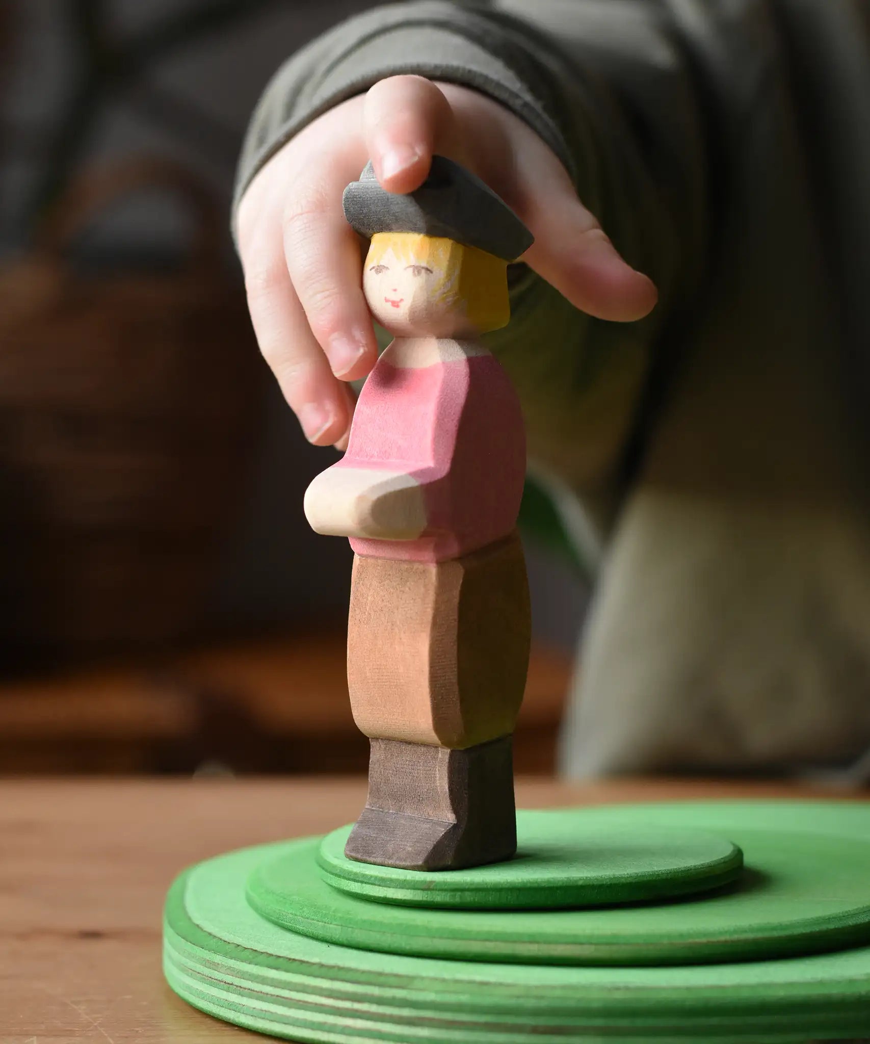 Child's hand reaching for the Ostheimer son wooden toy figure. The handmade boy figure is wearing a black hat, red top, brown trousers and brown boots.