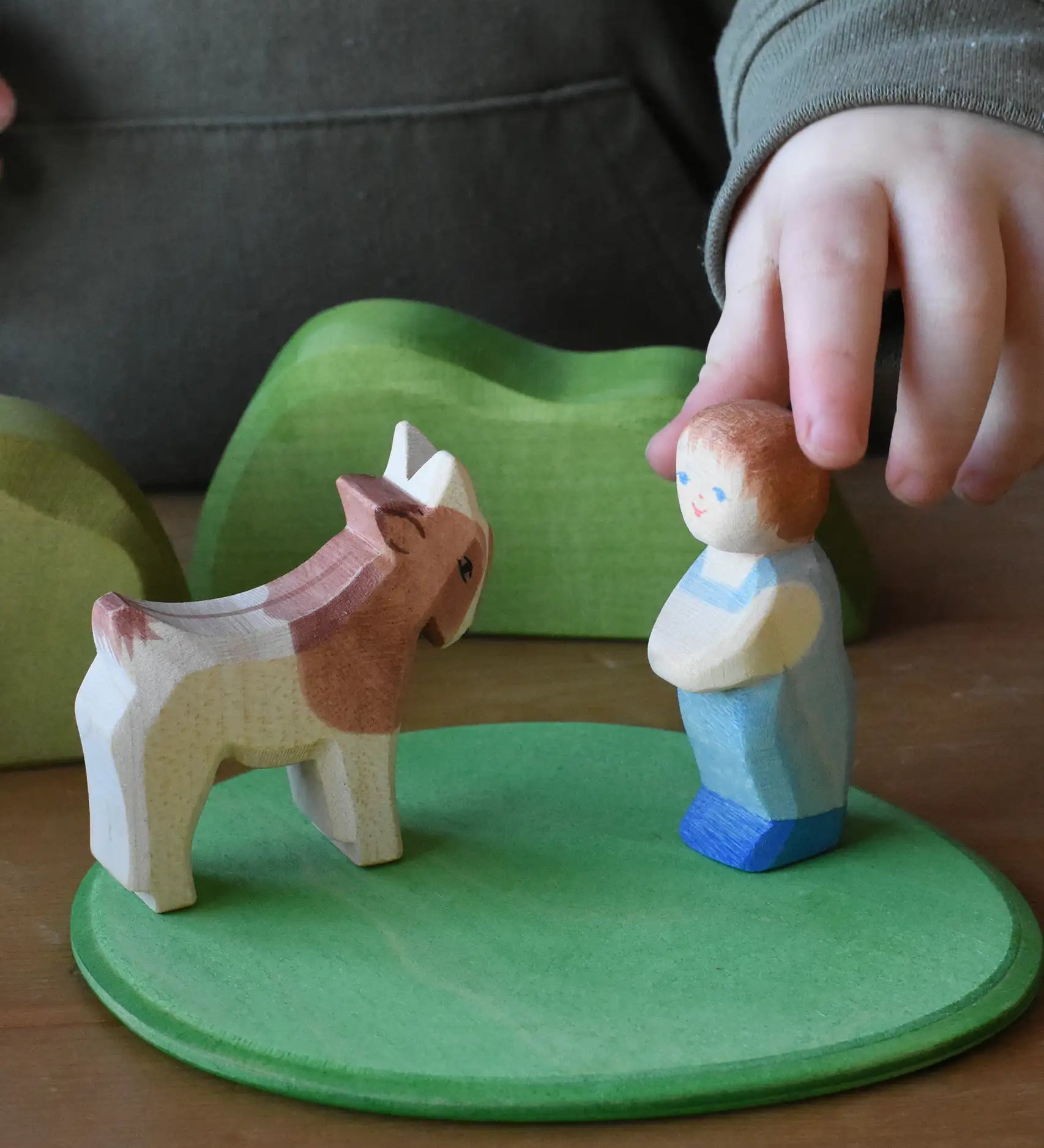 Child playing with the white skin toddler child wooden figure placing it on a green meadow play base piece next to a goat. 