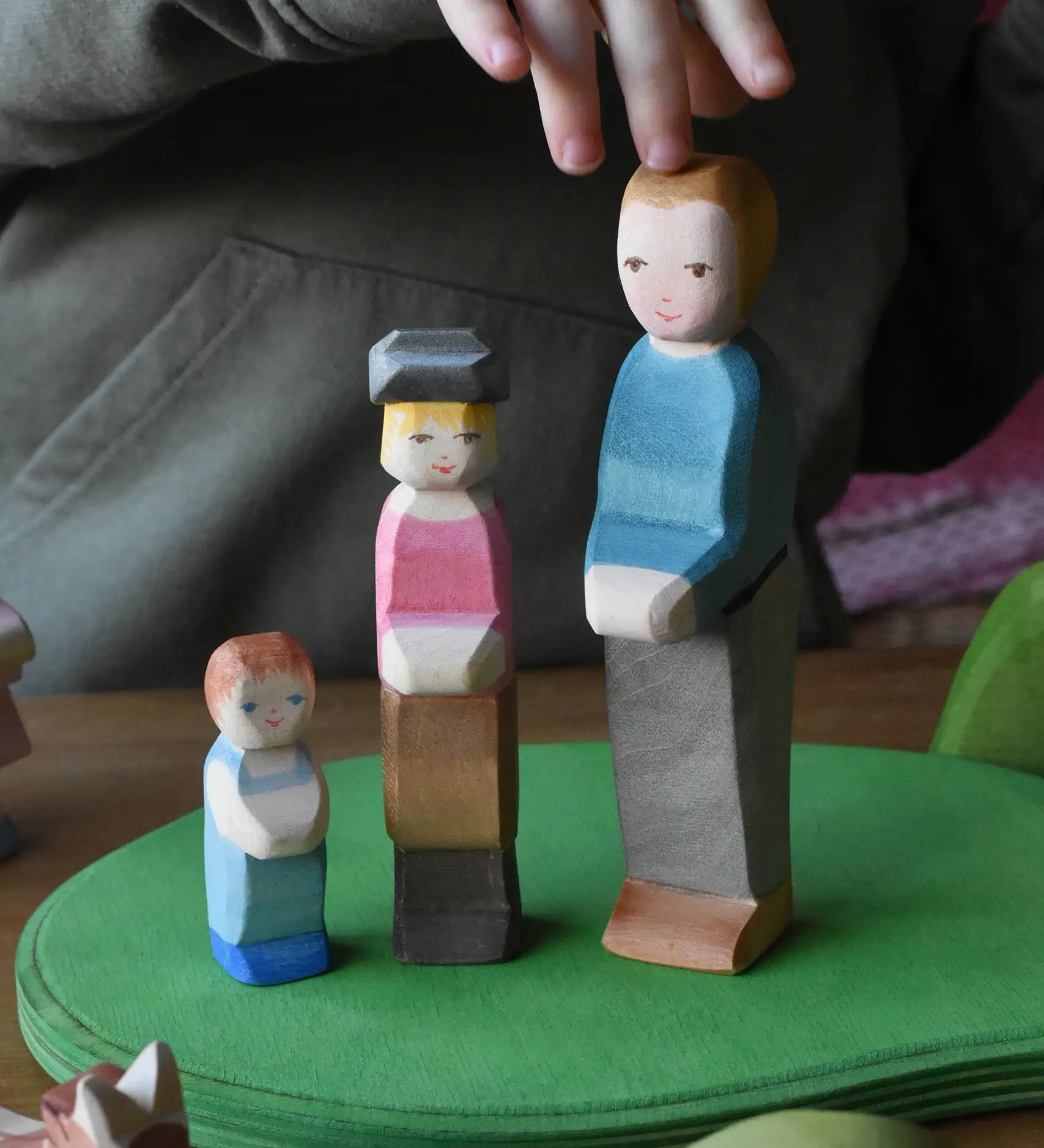 Child playing with Ostheimer wooden toy figures on a green meadow play base. A toddler child, son and a father figure are lined up next to each other.