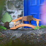 Ostheimer wild boar sow and piglet wooden toy figures placed on a wooden toy bridge in a small world play scene set up on a stone wall outdoors. Ostheimer green grass and an Oak tree can be seen off to the side of the scene.. 