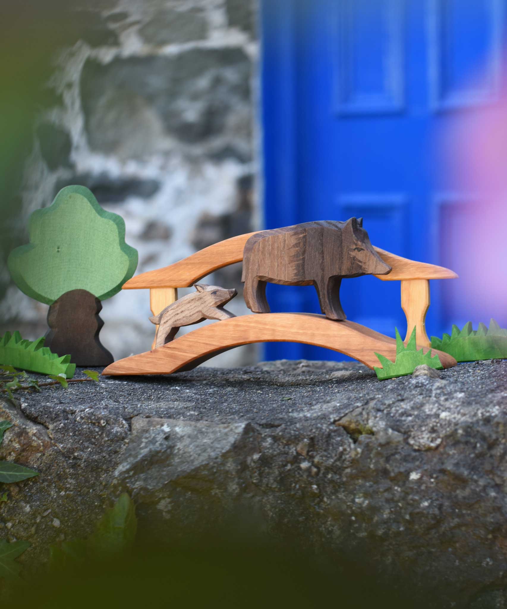 Ostheimer wild boar sow and piglet wooden toy figures placed on a wooden toy bridge in a small world play scene set up on a stone wall outdoors. Ostheimer green grass and an Oak tree can be seen off to the side of the scene.. 
