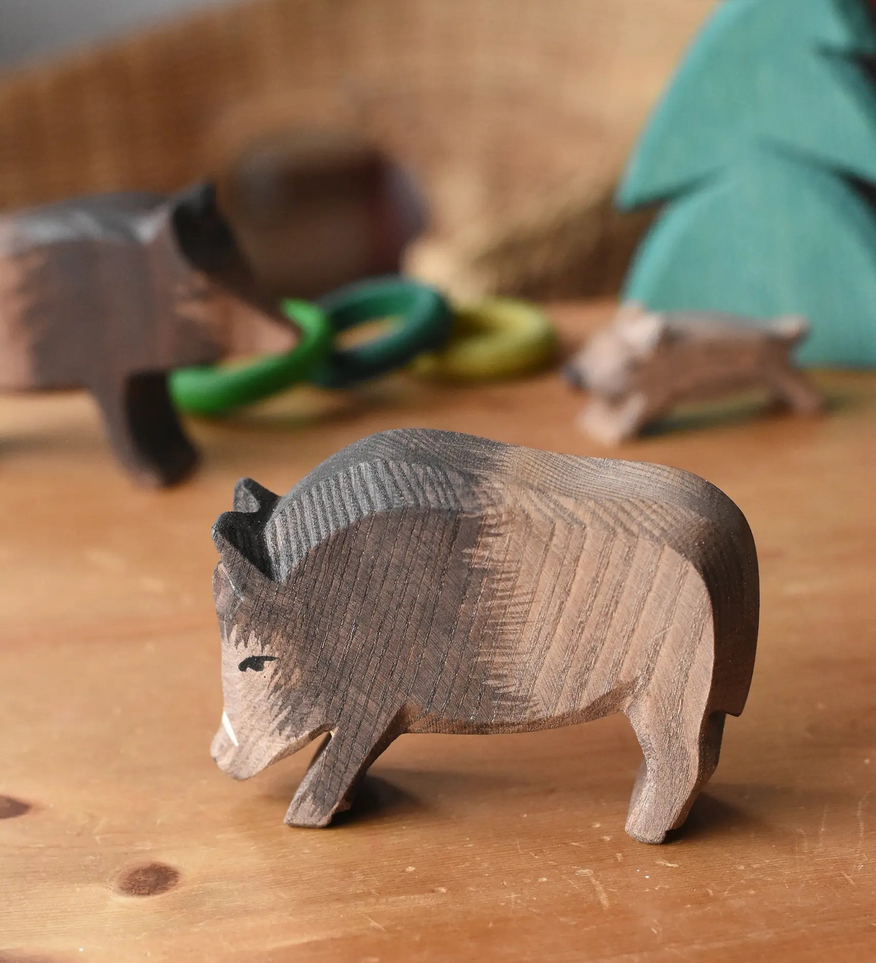 Ostheimer wild boar wooden toy figure placed on a wooden surface with other wild boars and a small spruce tree in the background. 
