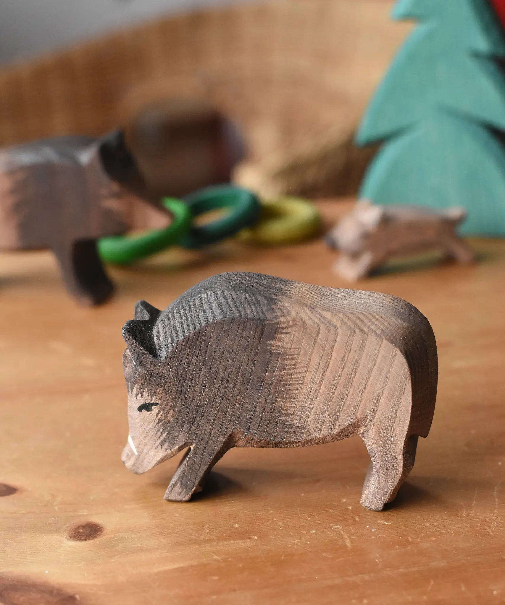 Ostheimer wild boar wooden toy figure placed on a wooden surface with other wild boars and a small spruce tree in the background. 