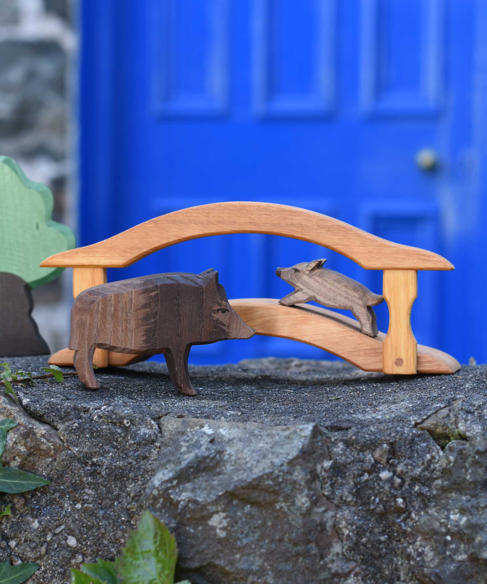 Ostheimer wild boar sow and piglet wooden toy figures placed on a wooden toy bridge in a small world play scene set up on a stone wall outdoors. Ostheimer green grass and an Oak tree can be seen off to the side of the scene.. 