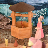 An Ostheimer hand made wooden wishing well toy placed on a wooden surface in a small world play scene with a lady in waiting figure besides it. These wooden figures are part of a wide range of expertly hand crafted wooden toys here at Babipur. 