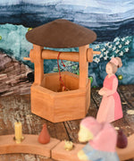 An Ostheimer hand made wooden wishing well toy placed on a wooden surface in a small world play scene with a lady in waiting figure besides it. These wooden figures are part of a wide range of expertly hand crafted wooden toys here at Babipur. 