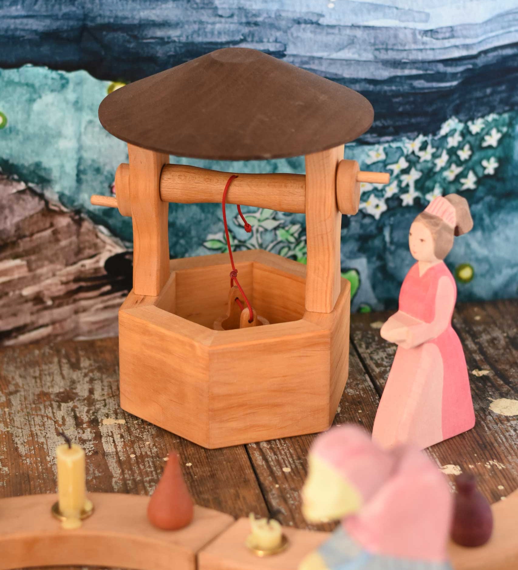 An Ostheimer hand made wooden wishing well toy placed on a wooden surface in a small world play scene with a lady in waiting figure besides it. These wooden figures are part of a wide range of expertly hand crafted wooden toys here at Babipur. 