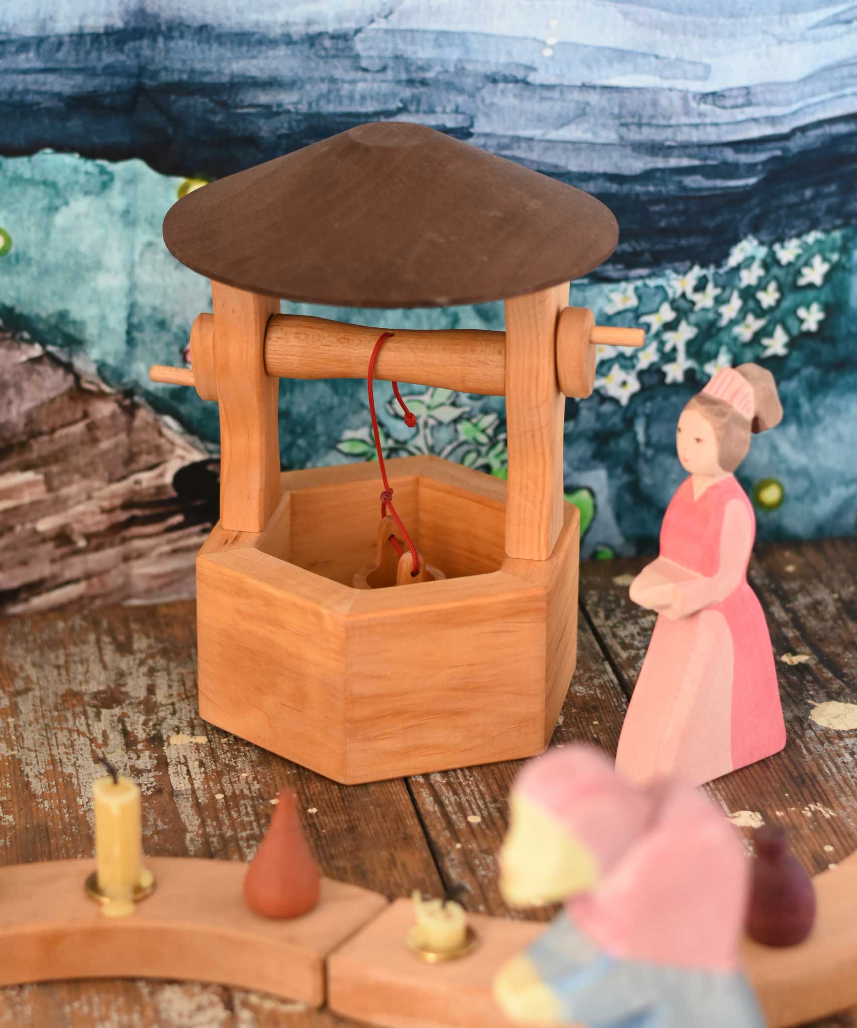 An Ostheimer hand made wooden wishing well toy placed on a wooden surface in a small world play scene with a lady in waiting figure besides it. These wooden figures are part of a wide range of expertly hand crafted wooden toys here at Babipur. 