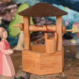 An Ostheimer hand made wooden wishing well toy placed on a wooden surface in a small world play scene with a lady in waiting figure besides it. Other Ostheimer wooden figures can be seen in the background. These wooden figures are part of a wide range of expertly hand crafted wooden toys here at Babipur. 