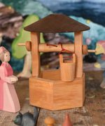 An Ostheimer hand made wooden wishing well toy placed on a wooden surface in a small world play scene with a lady in waiting figure besides it. Other Ostheimer wooden figures can be seen in the background. These wooden figures are part of a wide range of expertly hand crafted wooden toys here at Babipur. 