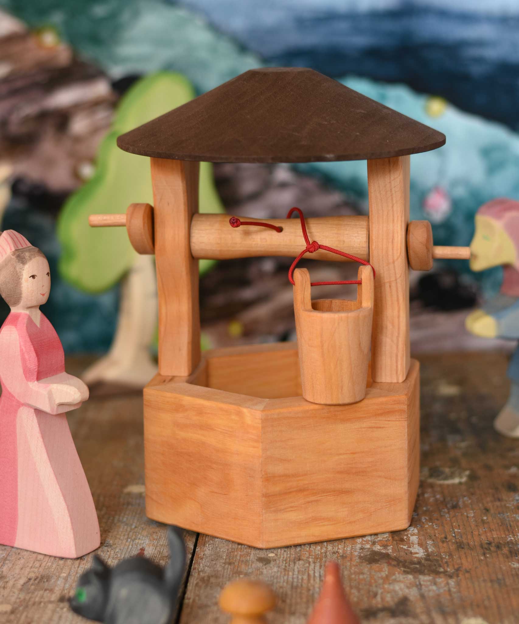 An Ostheimer hand made wooden wishing well toy placed on a wooden surface in a small world play scene with a lady in waiting figure besides it. Other Ostheimer wooden figures can be seen in the background. These wooden figures are part of a wide range of expertly hand crafted wooden toys here at Babipur. 