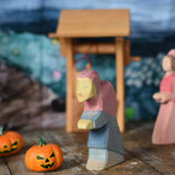 An Ostheimer hand made and hand painted wooden witch figure placed on a wooden surface in a small world play scene. An Ostheimer wooden well and other wooden toys including Bumbu pumpkins can be seen in the background. These wooden figures are part of a wide range of expertly hand crafted wooden toys here at Babipur.