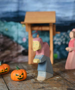 An Ostheimer hand made and hand painted wooden witch figure placed on a wooden surface in a small world play scene. An Ostheimer wooden well and other wooden toys including Bumbu pumpkins can be seen in the background. These wooden figures are part of a wide range of expertly hand crafted wooden toys here at Babipur.